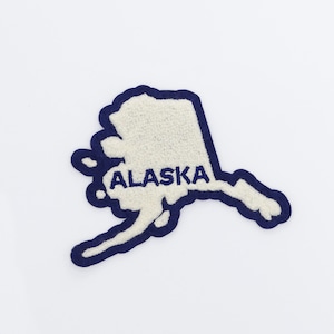 May include: A white and navy blue embroidered patch in the shape of the state of Alaska. The word "ALASKA" is embroidered in navy blue block letters within the patch.