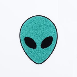 May include: A teal alien face patch with black oval eyes and a black border. The patch has a textured, fuzzy appearance and is set against a white background. The design is simple and graphic.