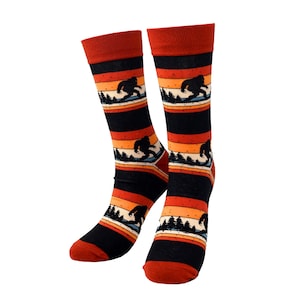 May include: A pair of novelty socks featuring a Bigfoot design. The socks have a black base with orange, yellow, and white stripes. Each sock has a Bigfoot silhouette and a forest scene. The toe and cuff are a solid rust color.