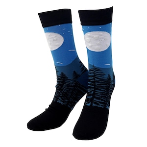 May include: A pair of crew socks with a night sky design. The socks feature a blue background with a white moon, stars, and a silhouette of a forest. The top and foot of the socks are black.