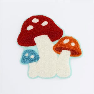 May include: A colorful, textured patch featuring three stylized mushrooms. The largest mushroom is red with white spots, the middle one is orange, and the smallest is blue. All have white stems and are outlined in a light blue.