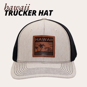 Puede incluir: Gorra de camionero beige y negra con un parche de cuero marrón con la palabra "Hawaii" y una escena tropical. La gorra tiene una parte trasera de malla y una visera curva. El texto "Hawaii Trucker Hat" está impreso en la parte superior.