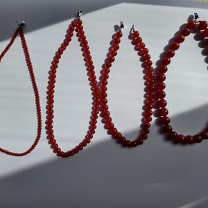May include: Four red beaded necklaces of varying lengths hang against a white background. The beads are a deep, rich red color. The necklaces are strung on a thin, silver-colored chain and are ready to wear.