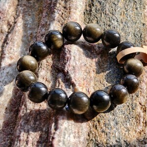 May include: A golden obsidian beaded bracelet. The bracelet is made of polished, round beads with a reflective, metallic sheen. The beads are strung together, forming a circular shape. A small tag with the text "RAW as." is attached.