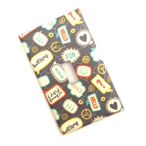 May include: A light switch cover with a gray background and a colorful pattern of speech bubbles with words like "hello", "fun", "awesome", and "cool".