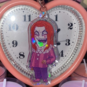 Könnte beinhalten: Ein holografischer Anhänger mit einer Comicfigur mit roten Haaren, einem rosa-violetten Outfit und einem verschmitzten Grinsen. Die Figur hält eine Hand auf der Hüfte und hat ein sternförmiges Design auf der Brust.