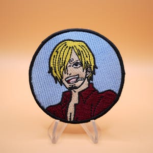 May include: A circular embroidered patch featuring a character with blonde hair, a dark red shirt, and a cigarette in their mouth. The character is set against a light blue background, and the entire patch is bordered by a thick black stitched edge.