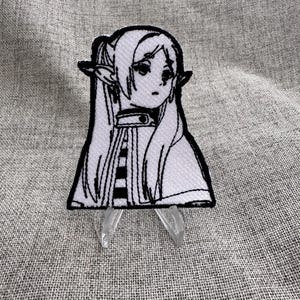 May include: A black and white embroidered patch featuring a manga-style female character with long hair and pointed ears. The patch is on a gray fabric background and is held up by a clear plastic stand.