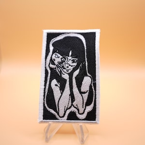 May include: A rectangular embroidered patch featuring a black and white graphic illustration of a person with long dark hair resting their chin on their hands, with a smaller face emerging from the side of their head, all bordered in white.