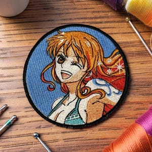May include: A circular embroidered patch featuring a stylized anime character with long orange hair, a winking expression, and a wide smile. The character is set against a blue background, framed by a thick black border, with white star accents.