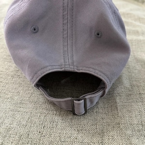 May include: A light purple baseball cap made of a soft fabric. The cap has a curved brim and an adjustable strap with a metal buckle. The cap has a classic six-panel design with a top button and stitched details.