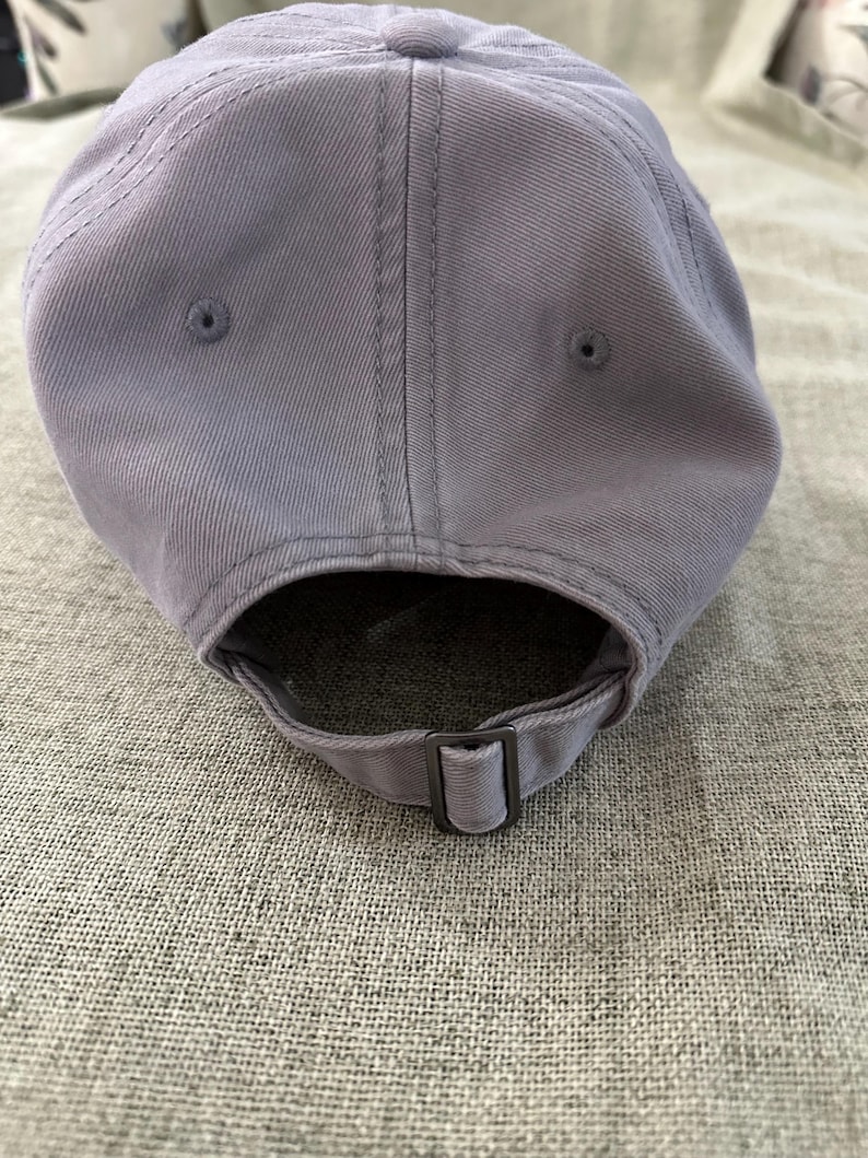 May include: A light purple baseball cap made of a soft fabric. The cap has a curved brim and an adjustable strap with a metal buckle. The cap has a classic six-panel design with a top button and stitched details.