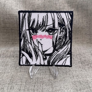May include: Square embroidered patch featuring a black and white anime-style illustration of a girl with pink blush on her cheeks. The patch has a black border and is displayed on a clear stand.