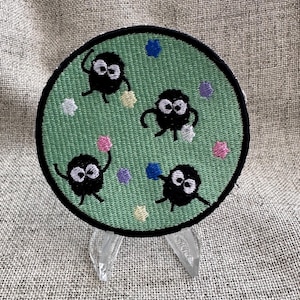 May include: A round, embroidered patch featuring four black, cartoon-like characters with white eyes and outstretched arms, set against a light green background. The patch is decorated with colorful star-shaped and round accents, and has a black border.