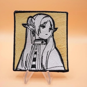 May include: A square embroidered patch featuring a stylized anime-style character with long hair and pointed ears. The design uses black thread for outlines on a light gray background, set against a tan square field with a thick black border.