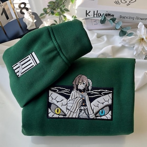 Puede incluir: Una sudadera verde con una imagen bordada de un personaje del anime "Demon Slayer". El personaje lleva una túnica blanca y una máscara. La sudadera también tiene un diseño bordado blanco en la manga.
