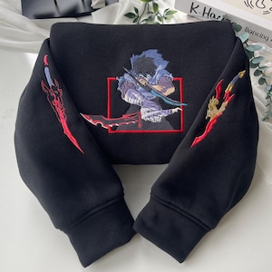 Op de afbeelding: Zwart sweatshirt met een rood geborduurd beeld van een personage dat een zwaard vasthoudt. Het personage bevindt zich in een rood kader. De mouwen hebben rode geborduurde ontwerpen.