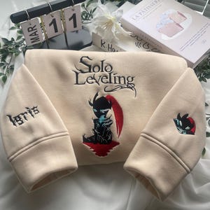 May include: A beige sweatshirt with a black embroidered design of a character from the "Solo Leveling" webtoon. The character is holding a sword and has red hair. The text "Solo Leveling" is embroidered in black above the character. The text "1871" is embroidered in black on the left sleeve.