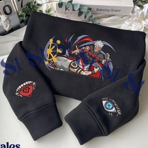 Op de afbeelding: Zwart sweatshirt met geborduurde anime-stijl kunstwerken. Het ontwerp toont een personage met rode en blauwe accenten en een geel wiel. Mouwen hebben geborduurde oog- en mondontwerpen. De merknaam "Stitchales" is zichtbaar.