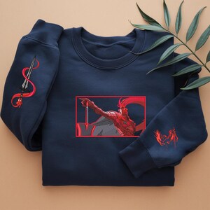 Puede incluir: Sudadera azul marino con bordados rojos. El dise&ntilde;o presenta una figura de guerrero con una espada y una lanza en la manga izquierda. La obra de arte est&aacute; enmarcada en un borde rojo. La sudadera es de material suave.