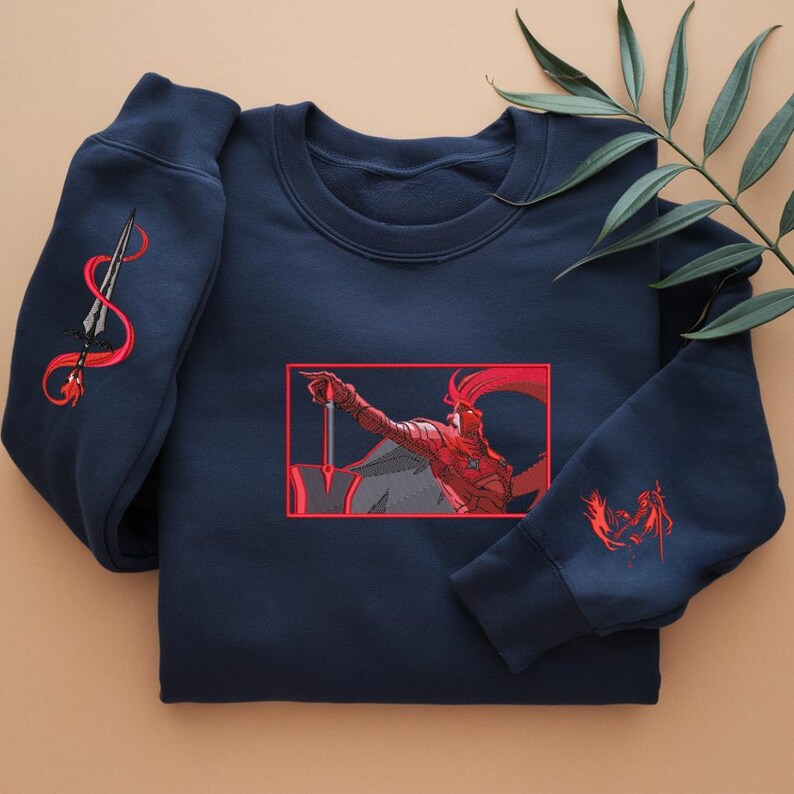 Puede incluir: Sudadera azul marino con bordados rojos. El dise&ntilde;o presenta una figura de guerrero con una espada y una lanza en la manga izquierda. La obra de arte est&aacute; enmarcada en un borde rojo. La sudadera es de material suave.