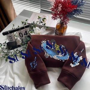 Puede incluir: Una sudadera marrón con un diseño bordado detallado que presenta personajes y olas. Las mangas tienen detalles bordados adicionales. La sudadera se muestra sobre una superficie blanca con elementos decorativos en el fondo.