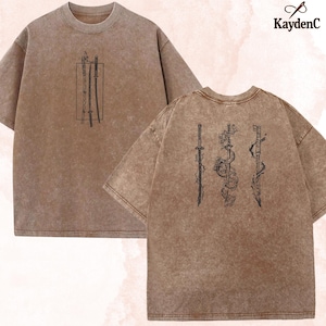以下が含まれることがあります： ブラウンのアシッドウォッシュ加工Tシャツ。フロントには長方形の枠内に3本の剣が描かれ、バックには龍や花のモチーフが施された3本の詳細な剣が描かれています。右上にKaydenCのブランド名がプリントされています。