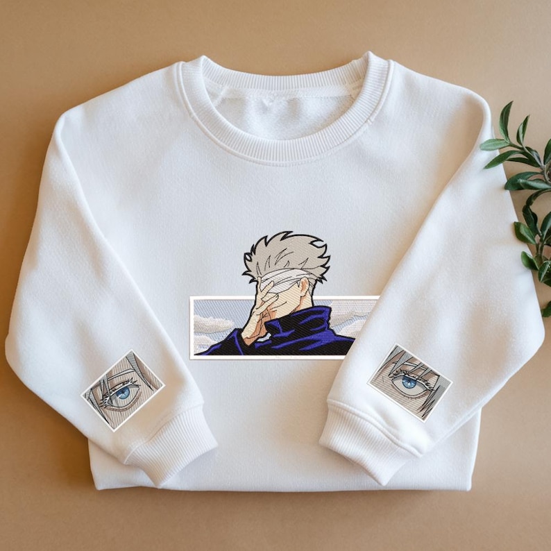 Pode incluir: Camisola branca com uma estampa gr&aacute;fica de um personagem com uma bandagem em um olho. O personagem est&aacute; vestindo uma gola alta azul e olhando para baixo. O gr&aacute;fico &eacute; cercado por uma borda branca. A camisola tem duas estampas gr&aacute;ficas menores do olho do personagem nas mangas.