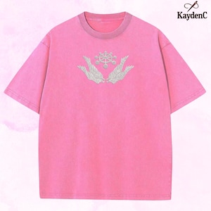 May include: A pink short-sleeved t-shirt with a crew neck and a faded look. The shirt has an embroidered design of two hands cradling a circular emblem with a detailed pattern. The brand name "KaydenC" is in the upper right corner.