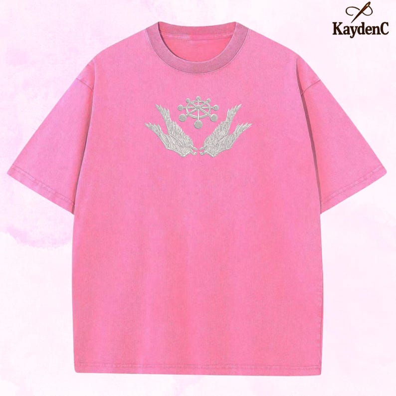 May include: A pink short-sleeved t-shirt with a crew neck and a faded look. The shirt has an embroidered design of two hands cradling a circular emblem with a detailed pattern. The brand name "KaydenC" is in the upper right corner.