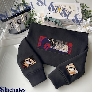 Puede incluir: Sudadera negra con gráficos bordados estilo anime. El diseño principal presenta un parche rectangular con una ilustración detallada. Gráficos de ojos adicionales están bordados en las mangas. La palabra "Stitchales" está impresa en la parte inferior.