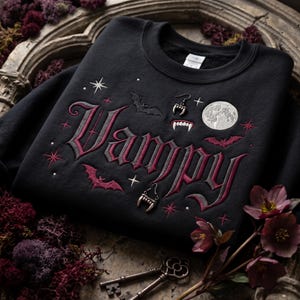 May include: A black crewneck sweatshirt featuring embroidered text that reads Vampy in a gothic font. The design includes embroidered details of a full moon, bats, and vampire fangs, accented with small red and white stars across the fabric.