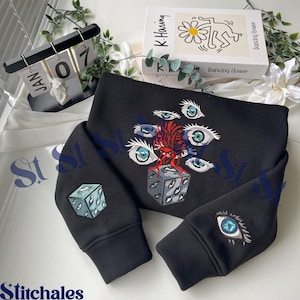 Könnte beinhalten: Schwarzes Sweatshirt mit gestickten Augen- und Würfeldesigns. Das Hauptdesign zeigt mehrere Augen und einen Würfel mit Augendetails. Die Ärmel haben einzelne Augenaufnäher. Im Hintergrund befindet sich ein Buch mit einer Keith-Haring-Illustration.