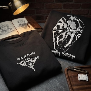 May include: Two black sweatshirts featuring white embroidered designs. One sweatshirt displays a graphic of a mouth with the text "King of Curses". The other sweatshirt features a large, detailed illustration of a character with multiple eyes and the text "Ryomen Sukuna".
