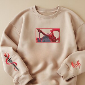 Puede incluir: Sudadera beige con ilustraciones bordadas. La parte delantera presenta una ilustraci&oacute;n roja y gris de una figura sosteniendo una espada dentro de un rect&aacute;ngulo rojo. La manga izquierda tiene un dise&ntilde;o de daga con detalles en rojo. La manga derecha tiene una ilustraci&oacute;n roja.