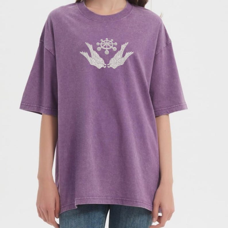 May include: A purple short-sleeved t-shirt with a crew neck. The shirt features a white graphic design of a stylized emblem with wings. The shirt has a slightly faded, vintage appearance.