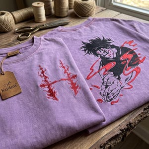 May include: A lavender t-shirt featuring a red embroidered flame-like letter H on the front and a large black and red embroidered anime-style character graphic on the back. A brown leather tag with the text KaydenC is attached to the collar.