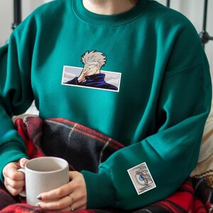 Pode incluir: Uma camisa de frio turquesa com um gr&aacute;fico de um personagem de anime branco e preto na frente e um gr&aacute;fico menor de um olho na manga.