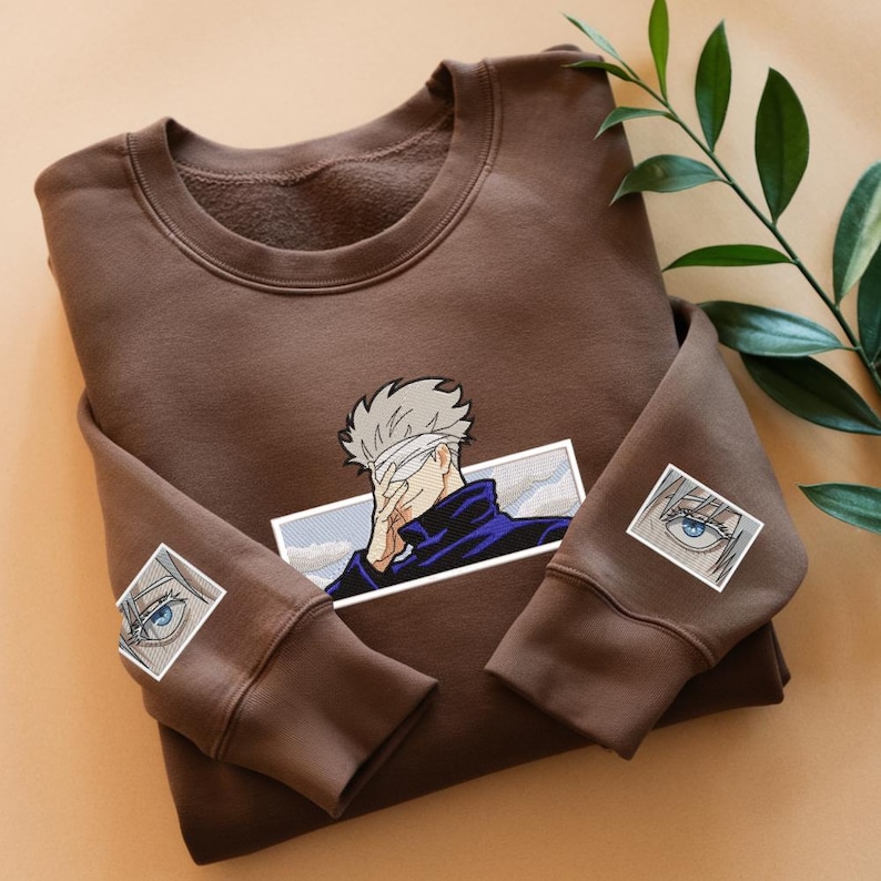 Pode incluir: Uma camisa de moletom marrom com uma imagem bordada em branco e preto de um personagem com uma venda nos olhos. O personagem est&aacute; vestindo uma camisa azul e est&aacute; cercado por uma nuvem branca. A camisa de moletom tem duas imagens bordadas menores de olhos nas mangas.