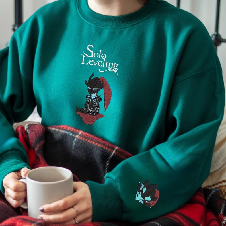 May include: A teal green sweatshirt with the text "Solo Leveling" and a black and red character illustration on the front. The character is wearing a helmet and holding a sword. The sweatshirt has a smaller version of the character on the left sleeve.
