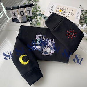 Puede incluir: Sudadera negra con diseño de personaje bordado estilo anime. Las mangas presentan una luna creciente amarilla y un sol rojo con una cruz. El diseño incluye detalles en azul y morado. La sudadera está doblada sobre una superficie blanca.