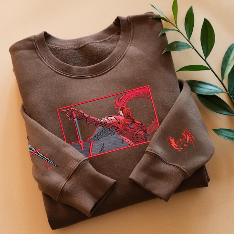 Puede incluir: Sudadera marr&oacute;n con cuello redondo y mangas largas. Presenta un gr&aacute;fico rojo y negro de un personaje con una espada en la parte delantera. La manga izquierda tiene un dise&ntilde;o de espada y la manga derecha un gr&aacute;fico rojo.