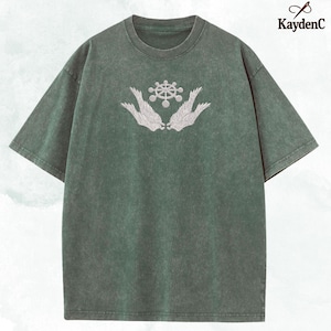May include: Green short-sleeved t-shirt with a crew neck. The shirt features a white embroidered design of a wheel-like shape above two stylized wings. The brand name "KaydenC" is printed in white on the upper right corner.