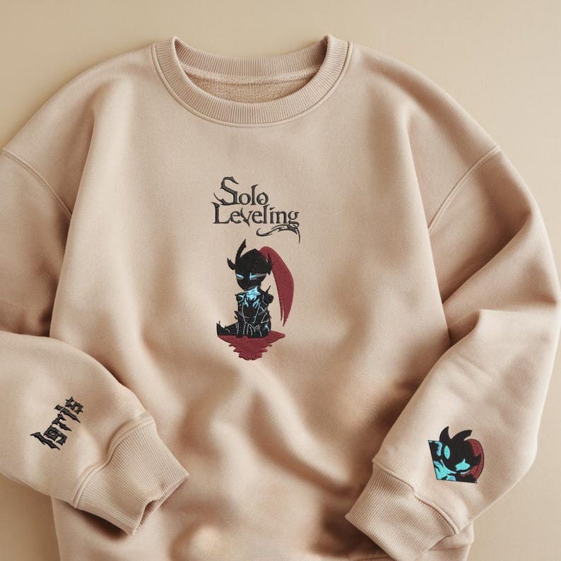 May include: A beige sweatshirt with the text "Solo Leveling" and a black and blue character illustration on the front. The left sleeve has the text "Solo" embroidered in black. The right sleeve has a black and blue character illustration.