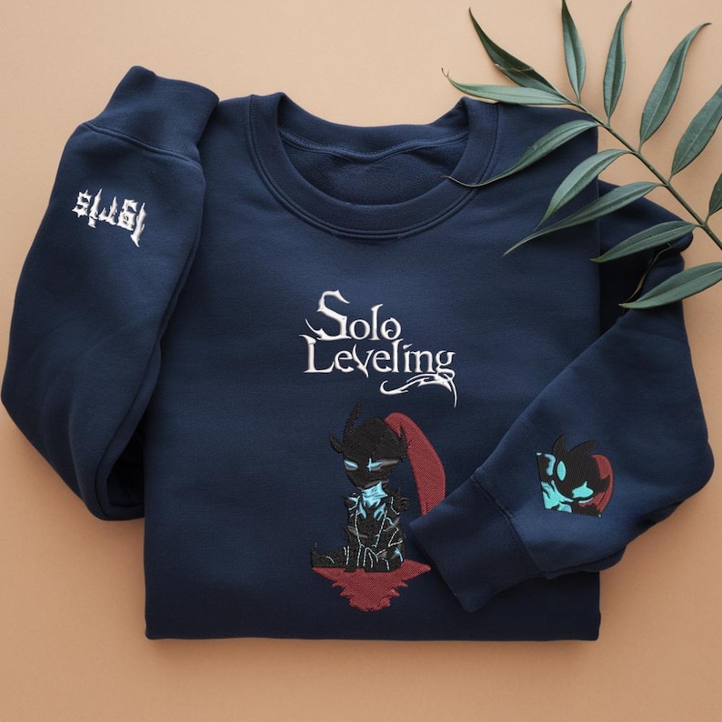 May include: A navy blue sweatshirt with the text "Solo Leveling" and an embroidered image of a character from the anime. The sweatshirt has a white embroidered logo on the left sleeve.