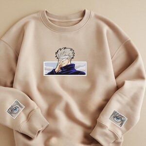 Pode incluir: Uma camisa de moletom bege com um personagem de desenho animado com os olhos vendados na frente e dois olhos de desenho animado menores nas mangas.