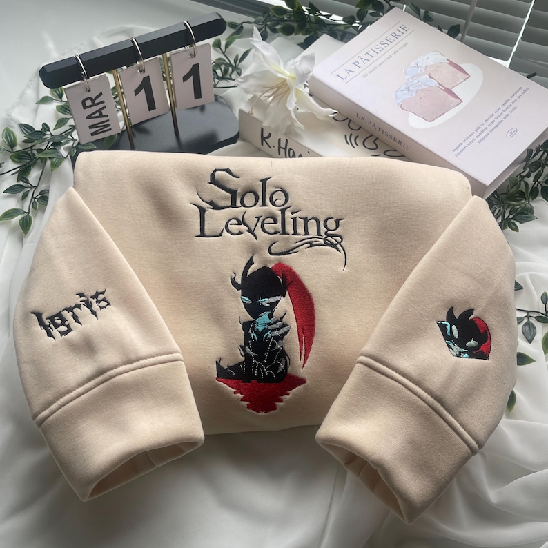 May include: A beige sweatshirt with a black embroidered design of a character from the "Solo Leveling" webtoon. The character is holding a sword and has red hair. The text "Solo Leveling" is embroidered in black above the character. The text "1871" is embroidered in black on the left sleeve.