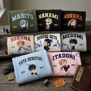 May include: A collection of eight folded sweatshirts in various colours, each featuring a different anime character illustration, a name in an arched font, and the text EST. 2017. The sweatshirts are displayed on wooden shelves with small spools of thread.