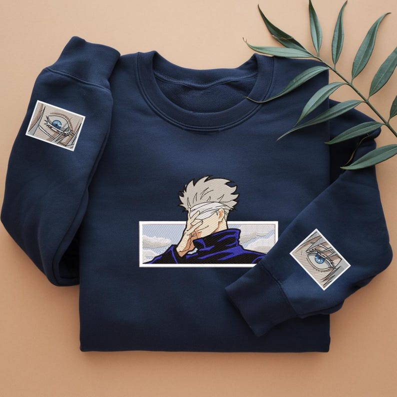 Pode incluir: Uma camisola azul-marinho com uma imagem bordada branca de uma personagem com uma venda nos olhos. A personagem est&aacute; a usar uma jaqueta azul e tem cabelo branco. A camisola tem dois olhos bordados brancos nas mangas.