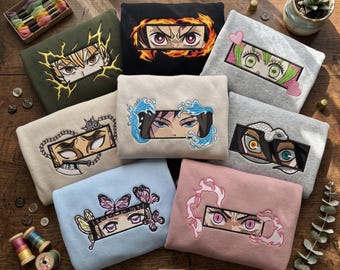 May include: A collection of eight folded sweatshirts in various colors, each featuring a rectangular embroidered patch displaying a pair of anime-style eyes surrounded by thematic elements like flames, water, butterflies, chains, and hearts.